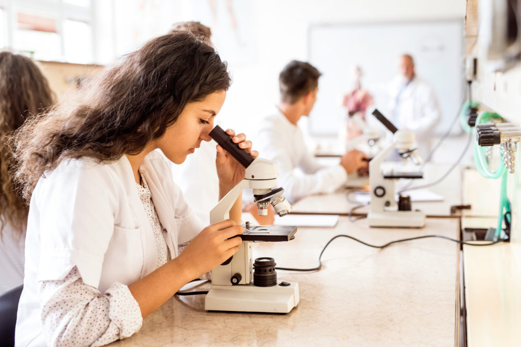 Beautiful hFemale college student with microscope in laboratory. Senior teacher teaching biology.