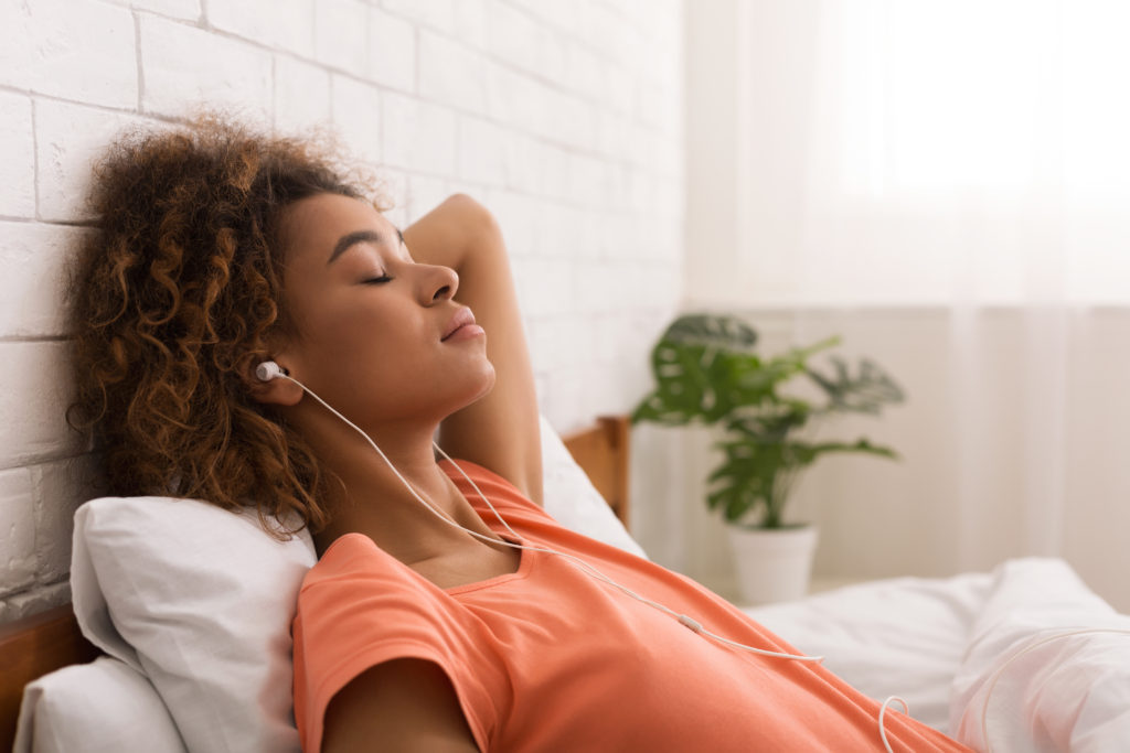 Young woman relaxing in bed with her eyes closed, wearing earbuds, and listening to ASMR videos to reduce her stress.